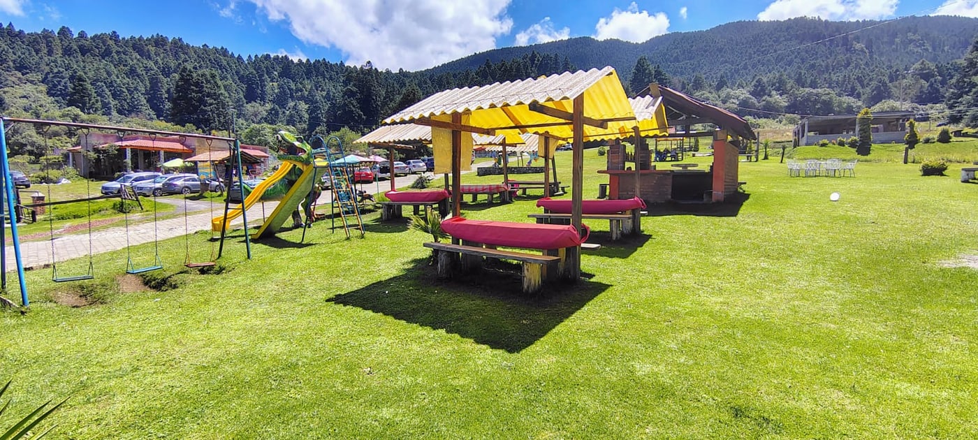 Palapa de techado amarillo con mesas sobre el área verde y la montaña al fondo